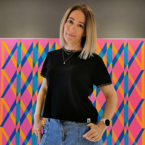 A woman wearing a black top and blue jeans is standing in front of a colourful, geometrically patterned painting.