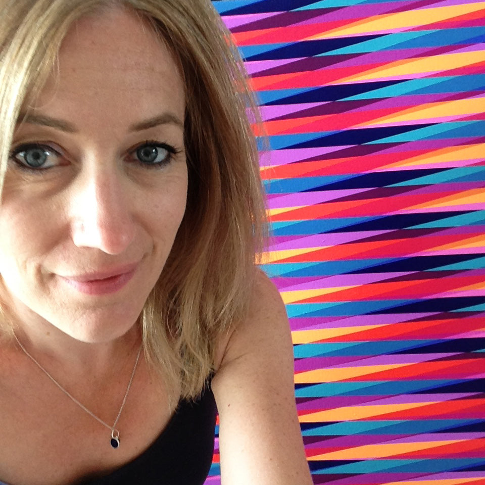 Portrait of artist Victoria Mardon, a woman with blonde hair and blue eyes, in front of a colourful striped abstract painting.
